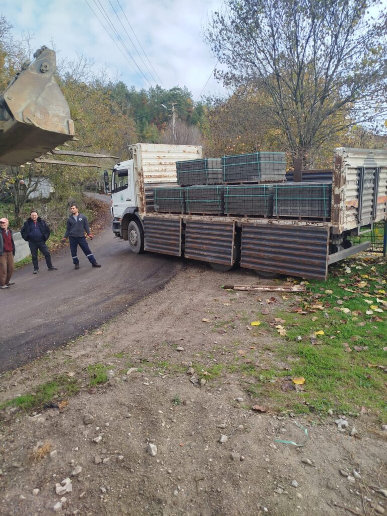 Satılmış Mahallesi’nde yol kenarına kaldırım taşı istifleyen bir kamyon, iş makinesi yardımıyla boşaltma yaparken, birkaç mahalle sakini süreci izliyor.